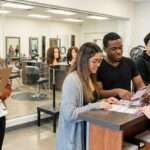 A diverse group of young adults standing at a modern beauty school reception desk, holding clipboards and brochures while discussing enrollment and program requirements with a friendly staff member.