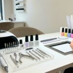 A detailed photograph of a professional nail technician's workstation, featuring essential tools for core procedures like an E-file, UV/LED lamp, precision steel implements, and product bottles. Hands are shown performing a sculpted nail extension using a form.
