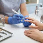 A close-up photograph of a professional, hygienic nail technology workstation. A technician in gloves uses a manicure pen on a client's natural nail matrix, positioned next to a sealed autoclave bag of sterile generic metal tools and a UV lamp on a pristine surface.
