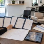 Prospective salon owner comparing blank state rule folders, permit packets, map outlines, salon chair samples, clean combs, a tablet, and keys on a large desk.