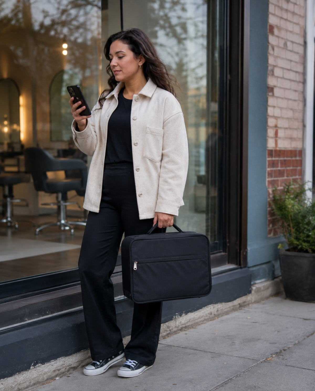 Cosmetologist standing outside an unmarked salon with a closed professional tool bag while checking her license application status on a phone