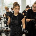 Cosmetology student blow-dries mannequin hair beside a blank monthly calendar in a classroom transitioning from daylight to evening.