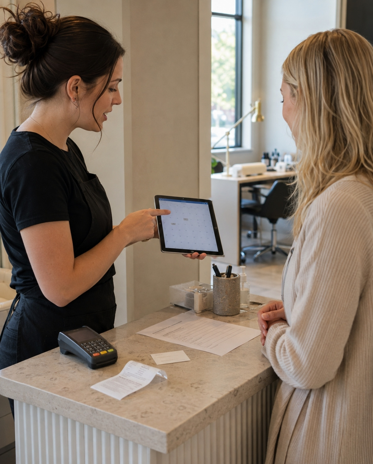 Nail technician explaining booking expectations to a client with a tablet calendar, deposit receipt, policy sheet, appointment card, and payment terminal visible.