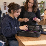 High school student opening a cosmetology training kit while an instructor explains a comb and hair sectioning clips.