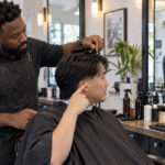 A barber lifts a client’s straight black top hair with a comb while the client points beside his ear to indicate his preferred fade height.